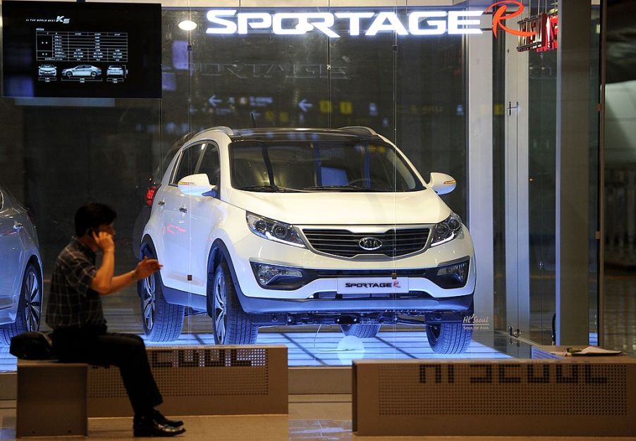 A man sitting next to a Kia Sportage in white on display. The most common Kia Sportage problems are quite disruptive.