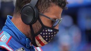 NASCAR driver Bubba Wallace stands with an American flag face mask, in his blue racing suit, with ear protection and sunglasses on