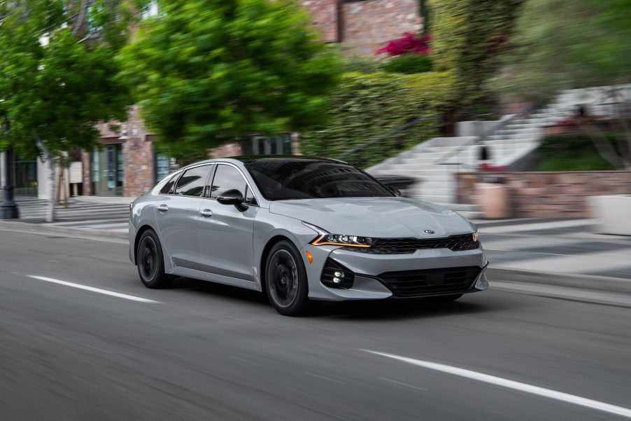 A gray 2021 Kia K5 sedan, successor to the Kia Optima, passes through a downtown city street.