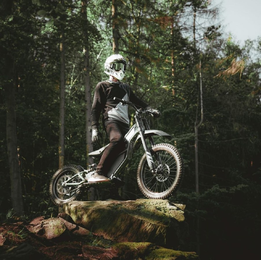 A white-helmeted rider on a green Kuberg Ranger, standing on a boulder in a forest