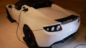 The rear of a white Tesla Roadster sits in a garage hooked up to a charging cable.