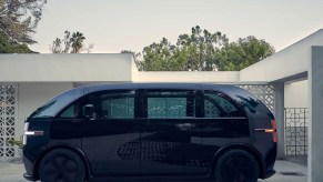 A black Canoo electric van sits in a driveway of a modern home.