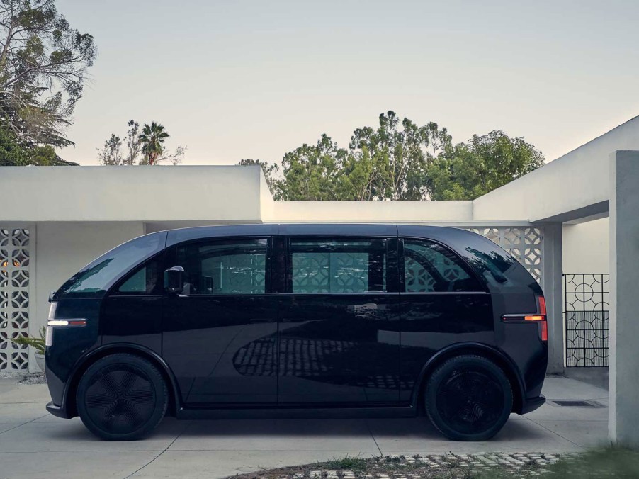 A black Canoo electric van sits in a driveway of a modern home.