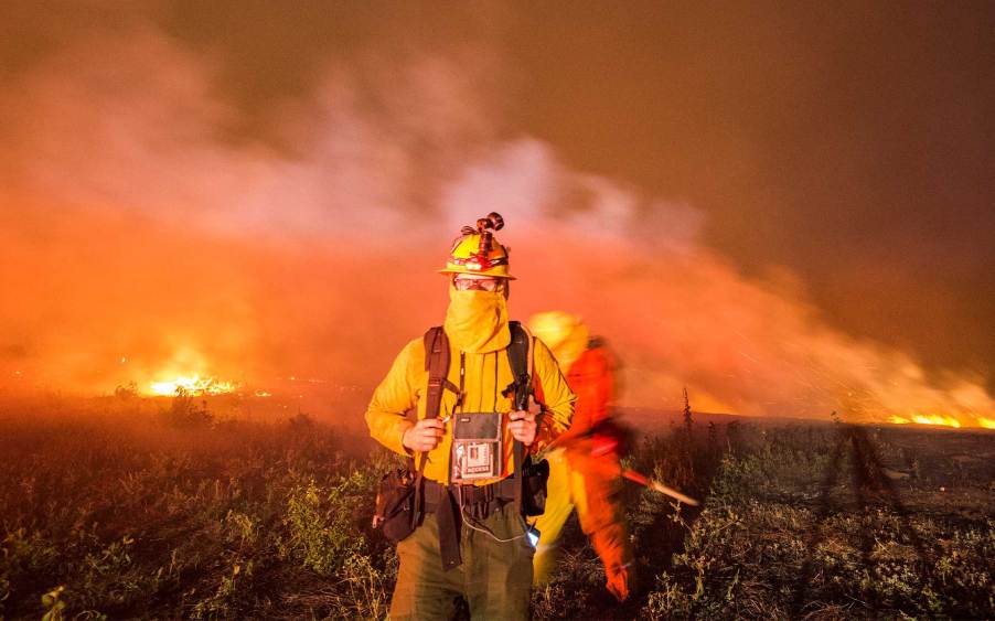 A firefighter in wildfire gear and camera in tow with a raging forest fire in the background