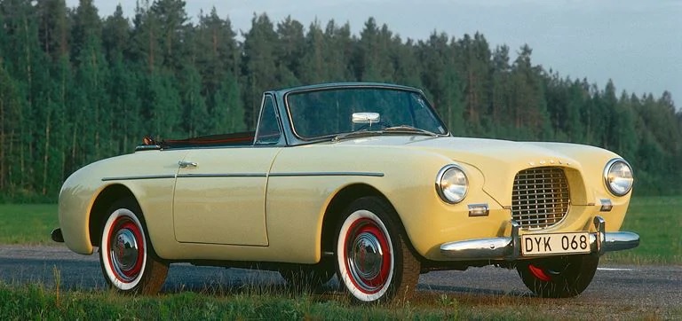 A yellow 1956 Volvo P1900 convertible in front of a forest