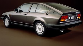 The rear-3/4 view of a gray 1980 Alfa Romeo GTV6 against a brown background