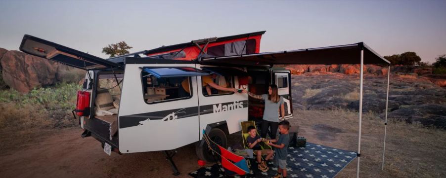 A white trailer RV with the pop-up extended sits on a trial with a family sitting under the exterior awning.