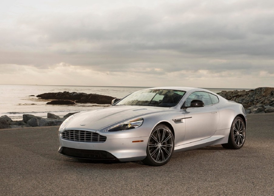 A silver Aston Martin DB9 sits on the side of a coastal road.