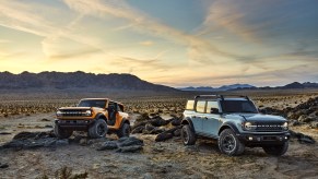 A pair of 2021 Ford Bronco SUVs parked on display over rugged terrain. One is a two-door model, and the other is a four-door model.