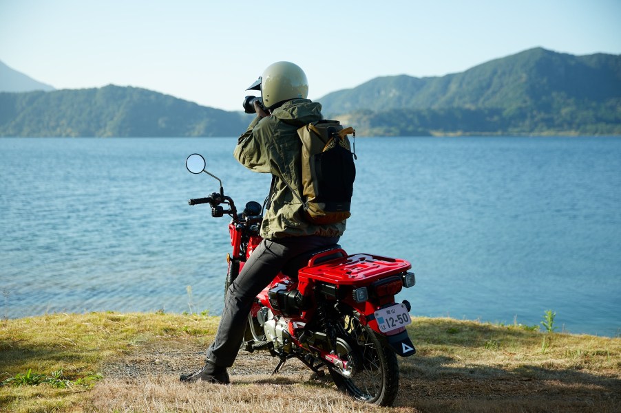 A rider sits on a red 2021 Honda Trail 125 by a lake