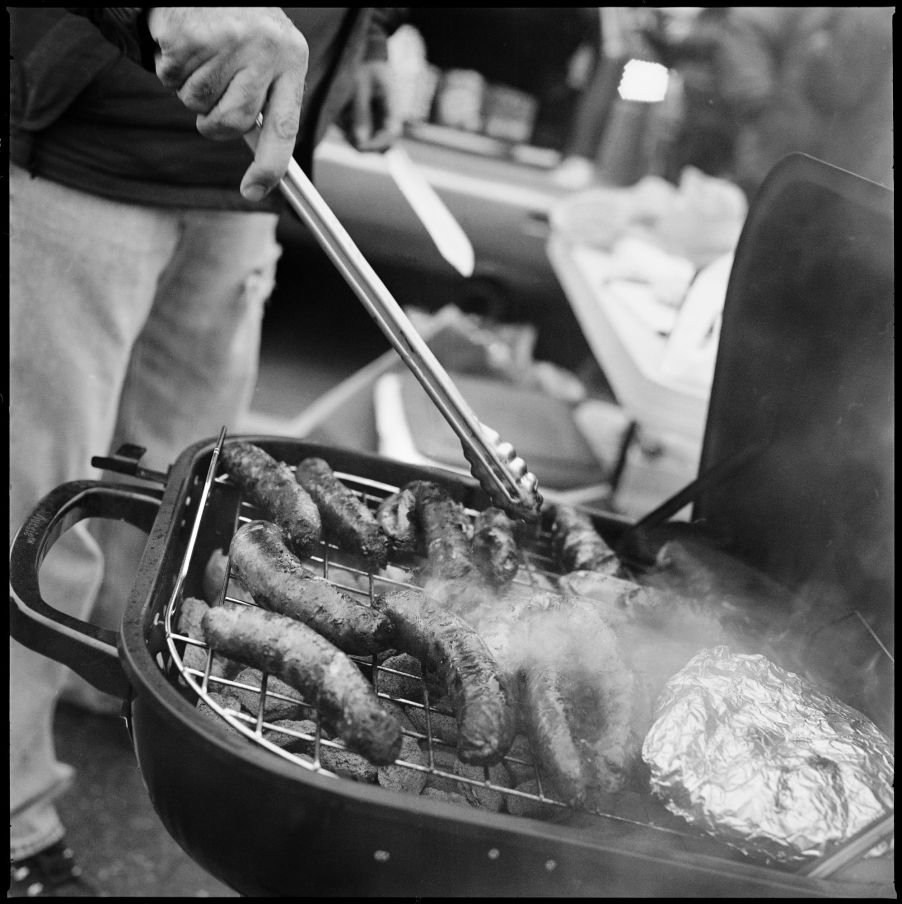 A tailgater cooks sausages on a grill by a car