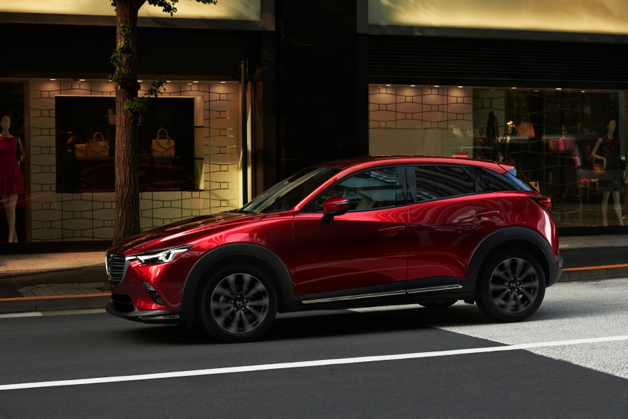 A metallic red 2019 Mazda CX-3 drives on a city street outside a storefront