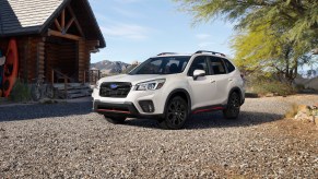 A White Subaru Forester is parked on gravel in front of a log cabin in the mountains.