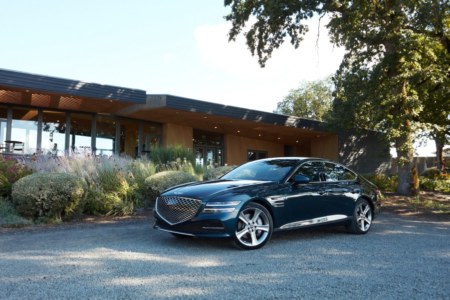 A dark-colored 2021 Genesis G80 parked in the shade of a tree outside a midcentury modern home