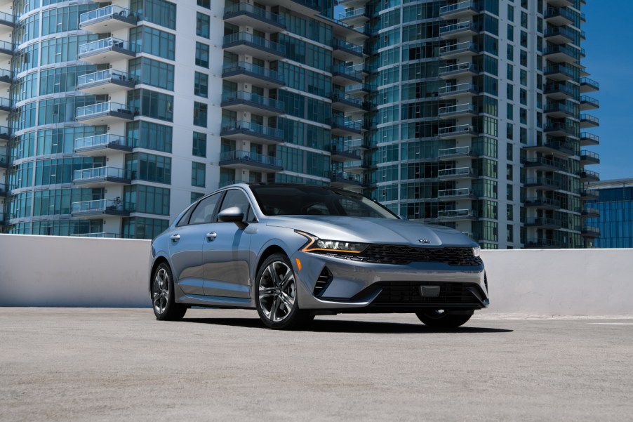 A silver 2021 Kia K5 parked on display with a skyscraper in the background