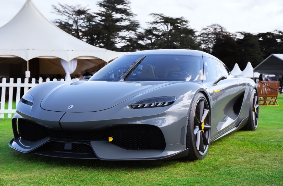 A gray Koenigsegg Gemera supercar is parked on a green lawn with a white tent in the background.