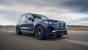 A dark-blue 2021 BMW Alpina XB7 large crossover SUV travels on a strip of asphalt surrounded by grassy hills