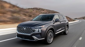 A dark-gray 2021 Hyundai Santa Fe travels on a two-lane highway flanked by hills on a cloudy day