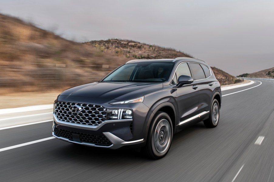 A dark-gray 2021 Hyundai Santa Fe travels on a two-lane highway flanked by hills on a cloudy day