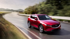 A metallic red 2021 Mazda CX-5 travels on a wet two-lane highway lined by metal guardrails and green mountains