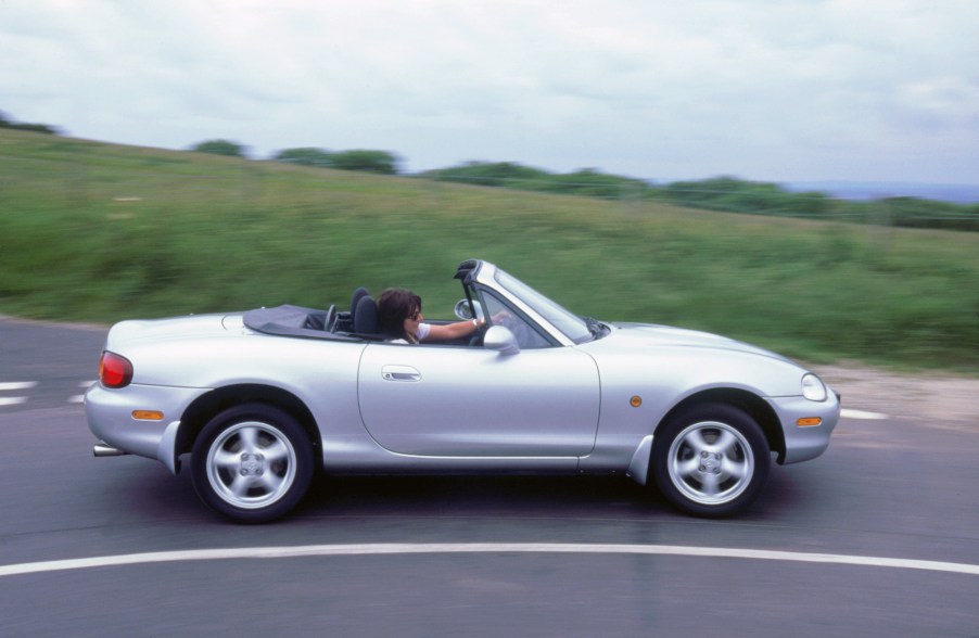 A person drives a silver 1999 Mazda MX5 on a curve in a road