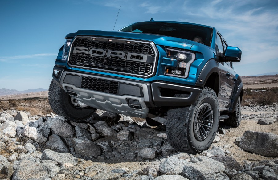 A bright-blue 2020 Ford F-150 Raptor parked on large rocks in a wide-open space with mountains and a blue sky in the background