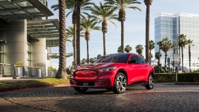 A red 2021 Ford Mustang Mach-E parked in a circular driveway outside a modern building lined with palm trees