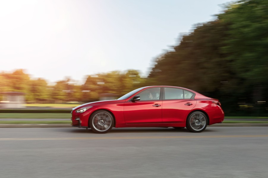 A red 2021 Infiniti Q50 sedan travels on a two-lane road alongside a park and trees