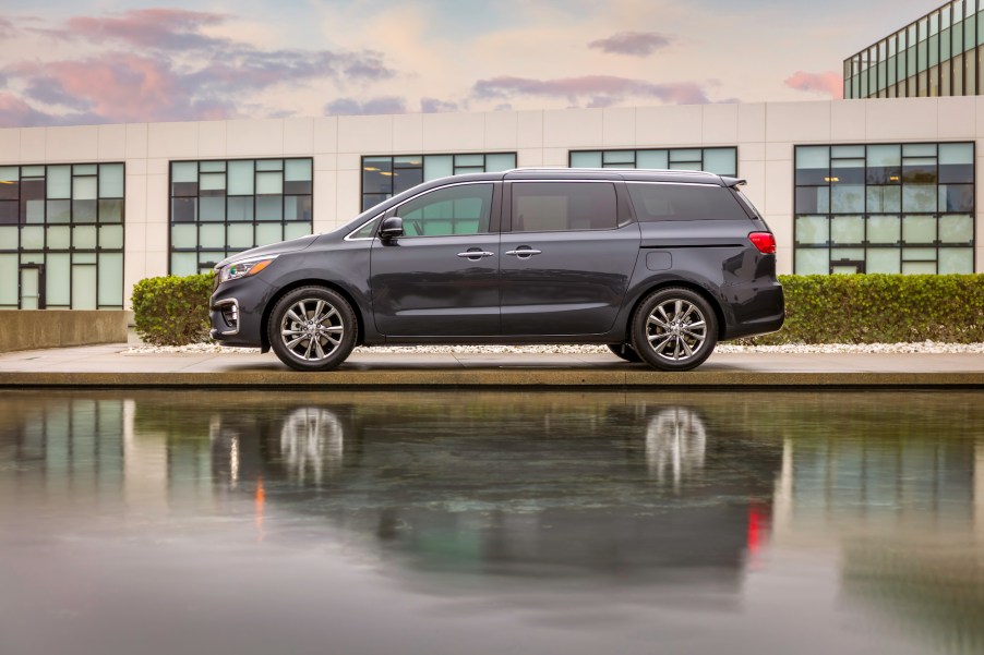 A dark-gray 2021 Kia Sedona parked between a body of water and a low-slung modern building