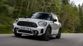 A white 2021 Mini Cooper Countryman travels on a road lined by pine trees on a cloudy day