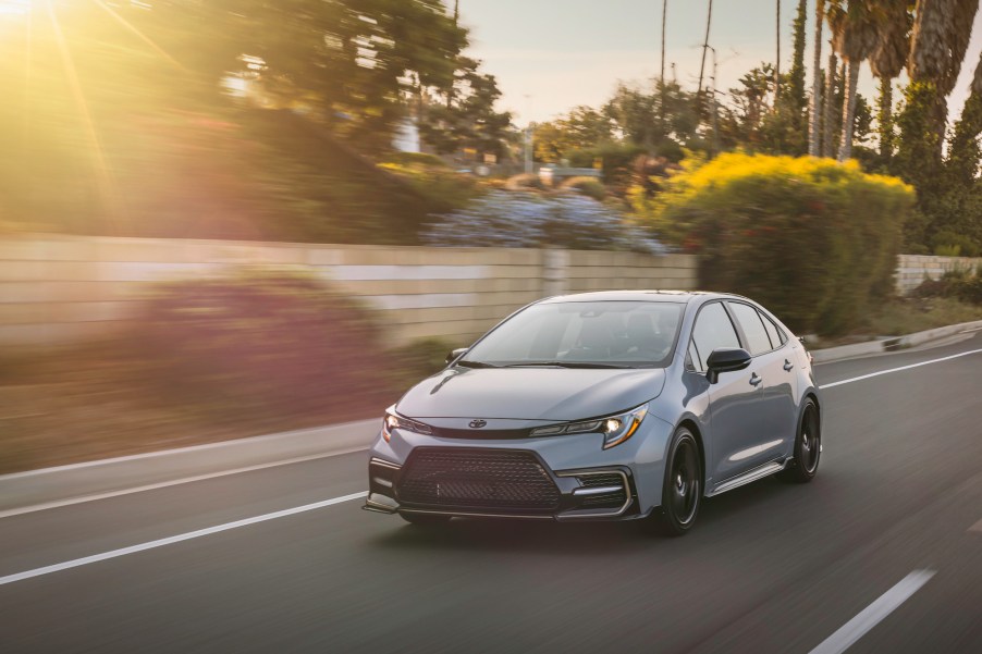 A silver 2021 Toyota Corolla Apex travels on a road along a wall and foliage