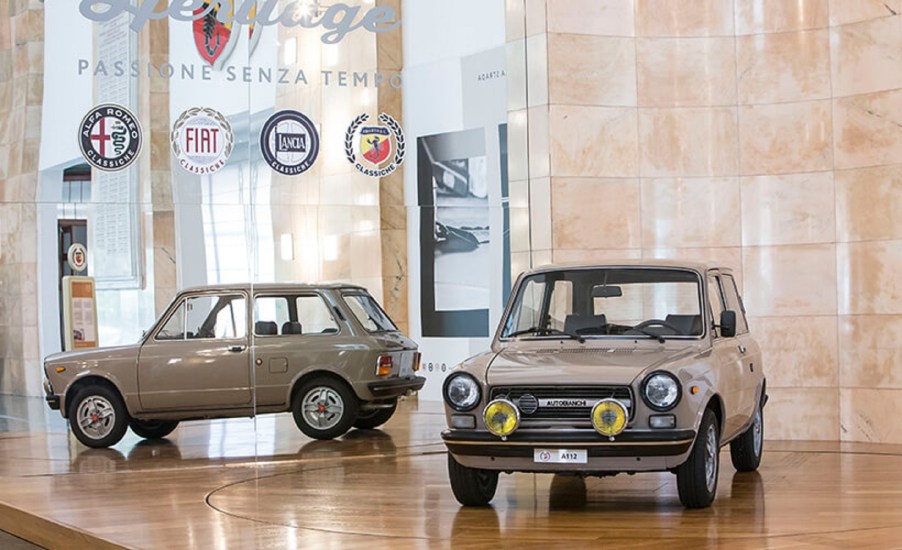 A brown 58-hp 1971 Autobianchi A112 Abarth in front on a mirrored Stellantis stand