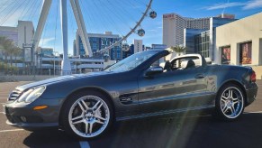 The side 3/4 view of a dark-gray 2005 Mercedes-Benz SL600 with its roof down by a Ferris wheel in a city parking lot