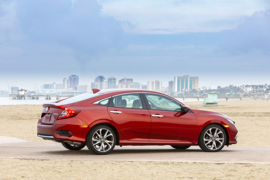 A red 2019 Honda Civic Sedan Touring compact sedan parked on a beach near a pier and city skyline on a cloudy day