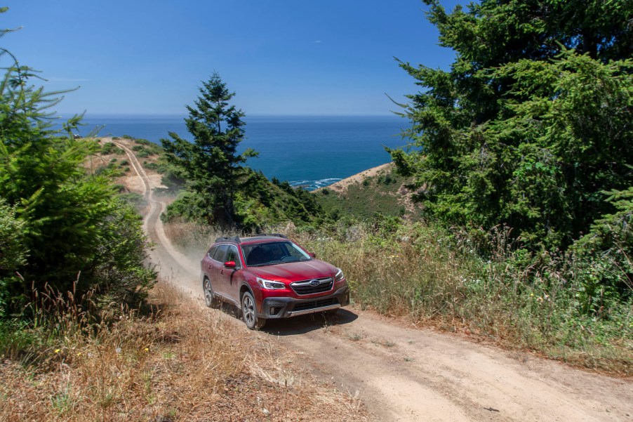 A red 2020 Subaru Outback midsize crossover SUV travels on a dirt trail up a pine-tree-dotted mountain with an ocean behind it