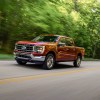 A metallic-red 2021 Ford F-150 Lariat four-door pickup truck traveling on a two-lane highway past thick trees on a sunny day