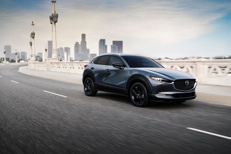 A metallic bluish-gray 2021 Mazda CX-30 2.5 Turbo subcompact crossover SUV travels on a bridge away from a city