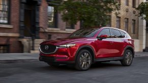 A red 2021 Mazda CX-5 driving down a city road with a building and tree in the background