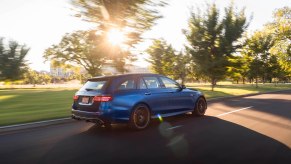 A blue 2021 Mercedes-AMG E63 S Wagon travels on a road lined with grass and trees on a sunny day