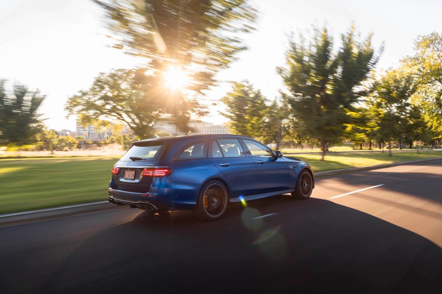 A blue 2021 Mercedes-AMG E63 S Wagon travels on a road lined with grass and trees on a sunny day