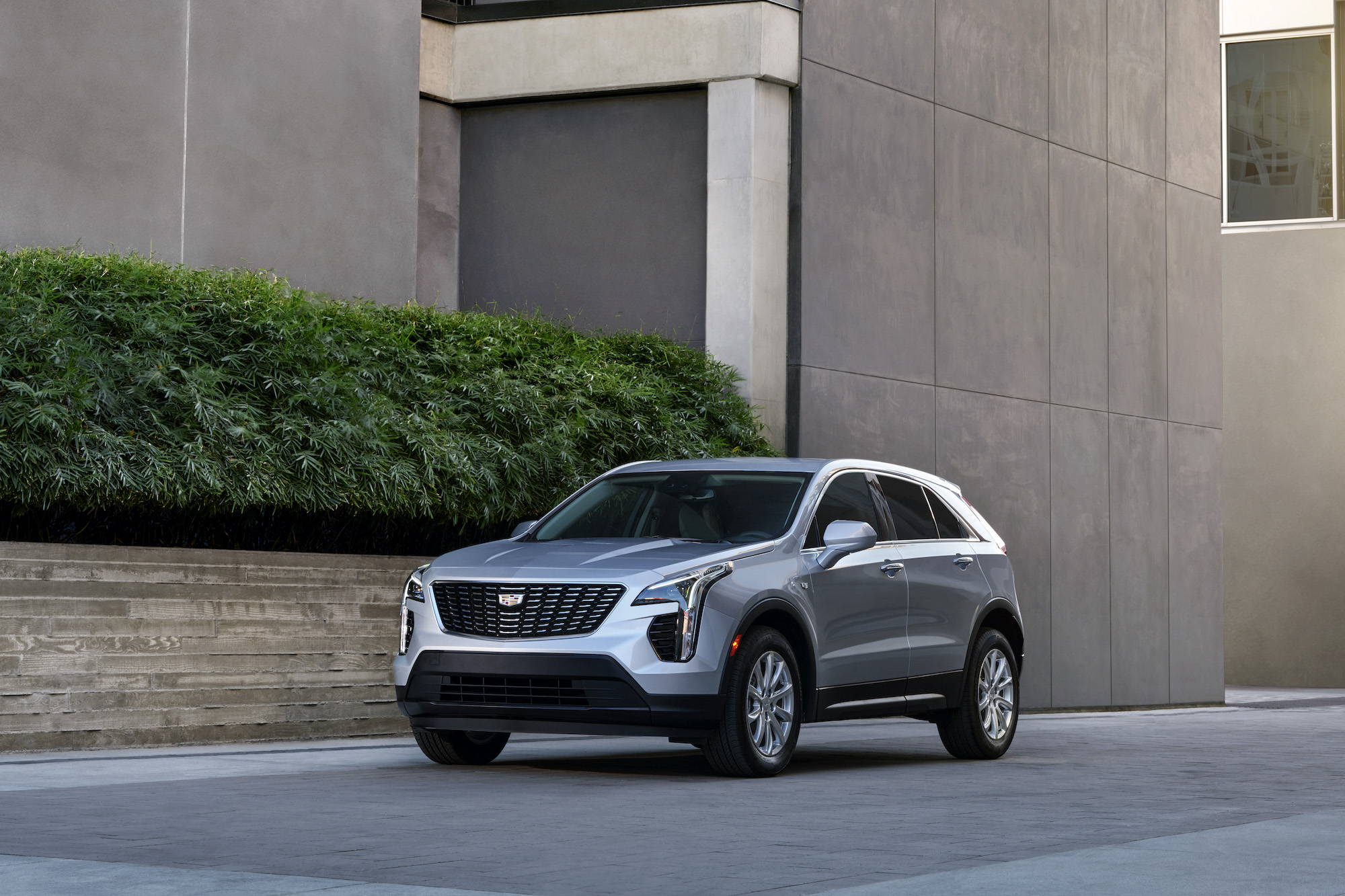 A silver 2021 Cadillac XT4 luxury compact crossover SUV parked next to a brownish-gray concrete modern building with a green hedge