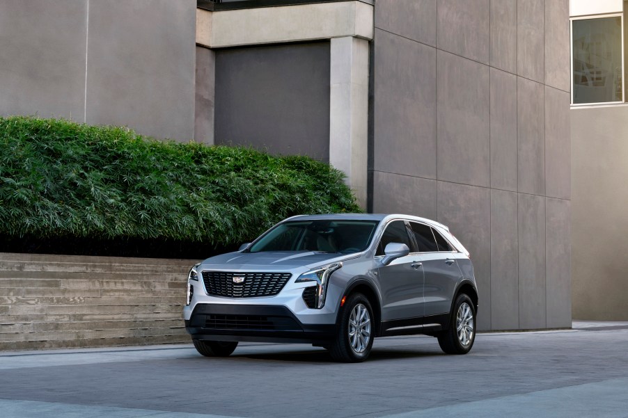 A silver 2021 Cadillac XT4 luxury compact crossover SUV parked next to a brownish-gray concrete modern building with a green hedge