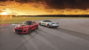 A red 2021 Chevy Camaro ZL1 coupe and a silver 2021 Chevy Camaro ZL1 convertible parked on a large expanse of asphalt overlooking grass, trees, and the sun on the horizon