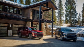 A red 2022 Jeep Wagoneer and a dark-colored 2022 Jeep Grand Wagoneer parked in front of a modern mountain home surrounded by snow and pine trees