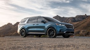 A blue 2022 Kia Carnival minivan parked on gravel in front of mountains on a partly cloudy day