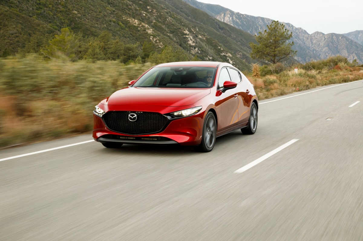 A red 2019 Mazda3 hatchback traveling on a highway past dry grass and mountains