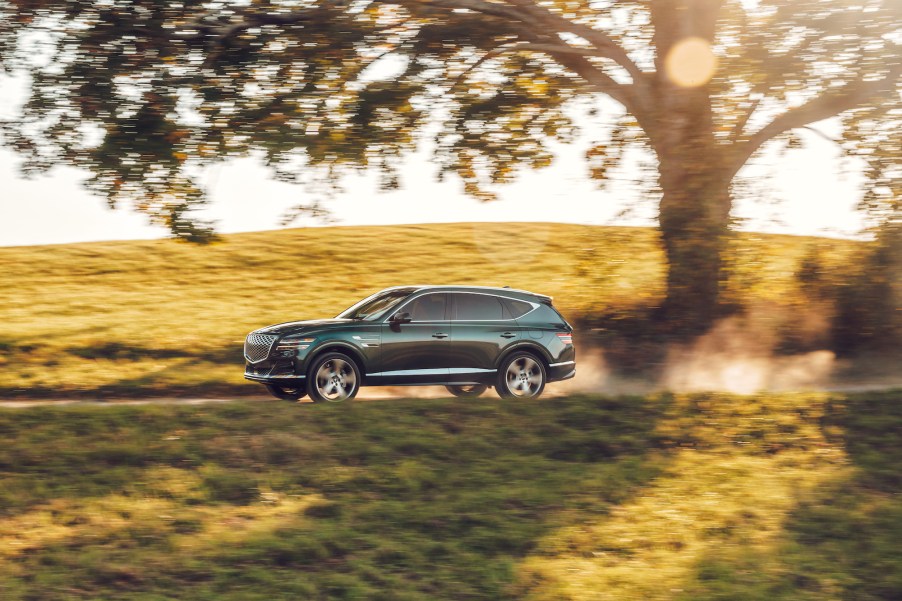 A dark-green 2021 Genesis GV80 midsize SUV traveling on a dusty road through rolling green hills in Hudson Valley, New York