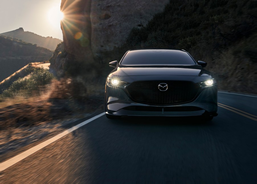 A dark-gray metallic 2021 Mazda3 2.5 Turbo compact car traveling on a two-lane highway next to a mountain