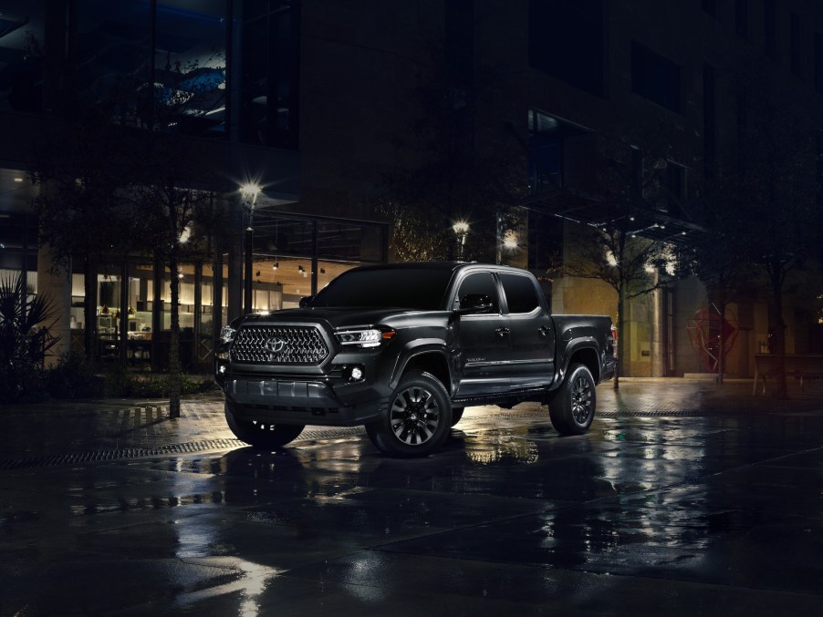 A black 2021 Toyota Tacoma Nighshade midsize pickup truck parked on wet pavement outside a glass building at night