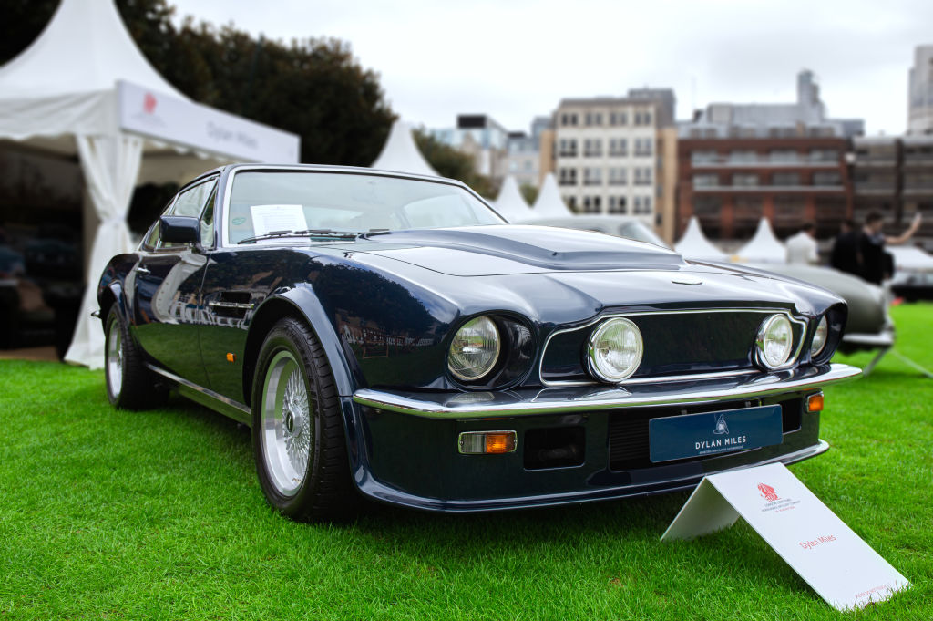 A dark blue 70s era Aston Martin V8 Vantage sits on a grass lawn 